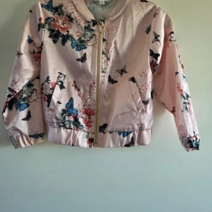 Blommig bomberjacka från KappAhl - Söt ljusrosa bomberjacka från KappAhl med blommigt och fjärilsmönster. Jackan har dragkedja i guld, ribbade muddar och två fickor framtill. Tillverkad i mjuk polyester med lite stretch för extra komfort. Perfekt för dig som gillar färg och mönster!