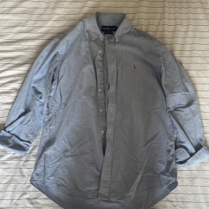 Blå skjorta från Ralph Lauren StL - Klassisk ljusblå skjorta från Ralph Lauren i 100% bomull. Custom fit-modell med button down-krage, broderad logga på bröstet och knäppning framtill. Har använts fåtal gånger men är i topp skick! 