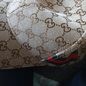 Äkta Gucci keps med GG-mönster och ränder -  Äkta Gucci. Snygg keps från Gucci i beige med klassiskt GG-monogram över hela kepsen. Har gröna och röda ränder på sidan och justerbar rem baktill i brunt läder. Tillverkad i bomullsmix och har en böjd skärm. Perfekt för dig som vill sticka ut med en lyxig accessoar.