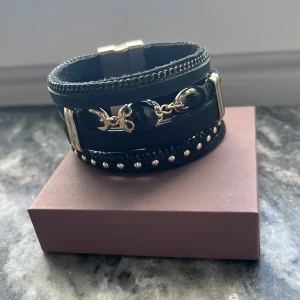 Svart och guldigt multiarmband - Snyggt svart armband med flera lager och detaljer i guld. Armbandet har en stor svart pärla i mitten, små guldfärgade nitar och kedjor som ger en cool och lyxig vibe. Magnetlås i silver gör det enkelt att ta på och av. Perfekt för dig som gillar statement-smycken. Köpt på second hand, så vet ej var det kommer från. 