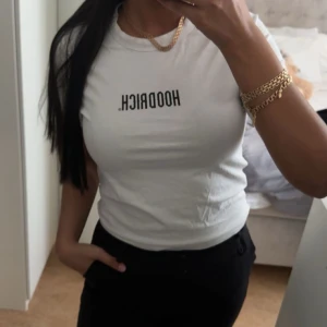 Vit t-shirt från Hoodrich - Vit t-shirt från Hoodrich med svart tryck på bröstet. Modellen är kortärmad och har en klassisk rund halsringning. Passformen är tight och materialet känns mjukt, troligtvis bomull. Perfekt för en clean och enkel streetstyle-look. Liten i storleken och är storlek XL men passar M 