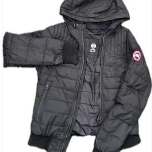 Svart cardigan Canada Goose - Svart cardigan Canada Goose med huva och klassisk logga på ärmen. Jackan är quiltad, har dragkedja framtill och ribbade muddar vid ärmslut och nederkant. Perfekt för kyliga dagar. Dock så finns de en deffekt på dragkedjan men kan såklart fixas om man vi lägga ner en extra slant! Storleken är S men passar även M.