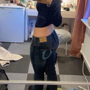 Mörkblå jeans med blå detalj - Säljer ett par mörkblå jeans med en stor blå broderad detalj på bakfickan. Jeansen har klassisk femficksdesign, raka ben och kontrastsömmar. Snygga och trendiga byxor som passar perfekt till en cool vardagslook.