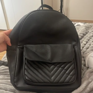 Svart ryggsäck från Primark i skinnimitation - Snygg svart ryggsäck från Primark i skinnimitation med quiltad ficka framtill och dragkedja upptill. Väskan har justerbara axelremmar och en praktisk innerficka med dragkedja. Perfekt för dig som vill ha en stilren och enkel look.