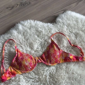 Färgglad bikiniöverdel med knyt - Snygg bikiniöverdel i rosa, orange och gul med abstrakt mönster. Den har bygel, smockade kupor och justerbara axelband. Baksidan har en söt knytning för extra detalj. Perfekt för strandhäng och poolparty!