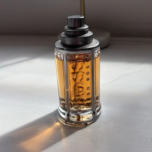 Hugo Boss The Scent parfym - Snygg parfymflaska från Hugo Boss The Scent med bärnstensfärgad vätska. Flaskan är cylindrisk med silverfärgade metallinslag och tydlig logga på framsidan. Modern och stilren design som känns lyxig och maskulin. Ca 45 ml kvar