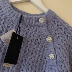 Ljuslila stickad kofta från YAS - Superfin ljuslila stickad kofta från YAS med glittriga dekorativa knappar vid axeln. Koftan har ett snyggt hålmönster, ribbade muddar och lång ärm. Perfekt för dig som vill ha en mysig men ändå stylish look.