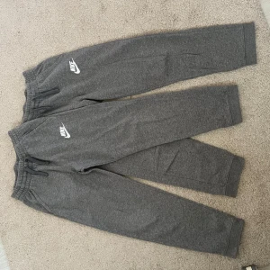 Grå mjukisbyxor från Nike  - Säljer ett par grå mjukisbyxor från Nike i juniorstorlek XL. Byxorna har elastisk midja med dragsko och Nike-logga i vitt på ena benet. Perfekta för chill eller träning, tillverkade i mjukt material som känns skönt mot huden! 