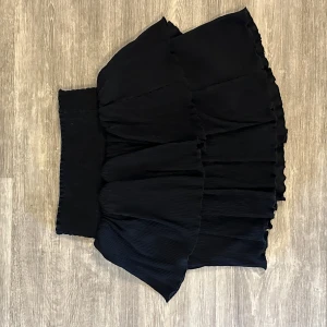 Svart volangkjol från Kappahl - Supersnygg svart kjol från Kappahl med flera lager volanger och bred, ribbad midja. Kjolen är kort och har en flowy känsla som ger extra rörelse. Perfekt för dig som gillar att sticka ut med en cool och feminin stil.