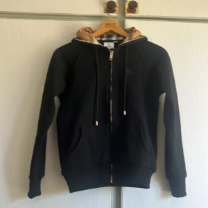 Svart hoodie från Burberry med dragkedja - Svart hoodie från Burberry med klassisk rutmönstrad huva i beige och rött. Tröjan har dragkedja framtill, snörning vid huvan och två fickor. Perfekt för dig som gillar stilrena streetwear-plagg med lyxig touch. OBS tröjan är mindre än S då den såldes till mig så!