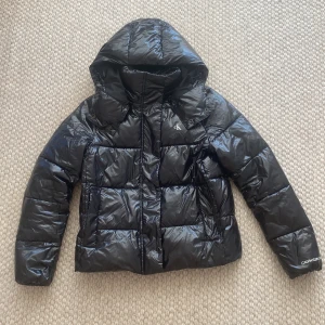 Svart pufferjacka från Calvin Klein - Säljer en svart, glansig pufferjacka från Calvin Klein Jeans. Jackan har huva, dragkedja framtill och diskret CK-logga på bröstet samt Calvin Klein Jeans-tryck på ärmen och insidan. Perfekt för kalla dagar och en riktigt snygg höst och vinter jacka💗💗