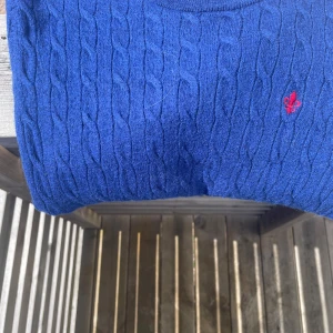  Morris tröja - Snygg blå kabelstickad tröja från Morris i storlek L, tillverkad i 100% merinoull. Tröjan har ett broderat rött emblem på bröstet och rund hals. Perfekt för dig som gillar klassisk stil med en twist och vill ha något mjukt och värmande. Riktig skön och perfekt för back too school för att få mer komplimanger 