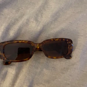 Rektangulära solglasögon från mango - Snygga solglasögon med rektangulära bågar i klassiskt leopardmönster. Bågarna är i plast med bruna och gula toner, vilket ger en cool vintagekänsla. Perfekta för att lyfta din stil under soliga dagar.