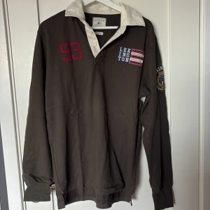 Brun/ typ grön  pikétröja Lexington med tryck - Snygg brun/ grön långärmad pikétröja från Lexington med vit krage, broderad siffra 93 och flaggdetalj på bröstet samt patch på ärmen. Klassisk rugbykänsla och skön bomullskvalitet. Perfekt för dig som gillar preppy stil.