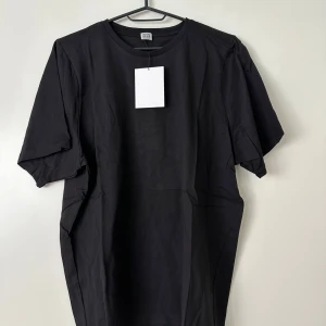 Svart t-shirt från Toteme - En stilren svart t-shirt från Toteme i 100% ekologisk bomull. Klassisk rund halsringning och avslappnad passform med korta ärmar. Perfekt basplagg för dig som gillar enkel och clean stil. Bröstvidd 55 cm, längd 65 cm, axelbredd 38 cm