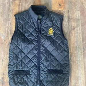 Svart quiltad dunväst från Morris - Snygg svart quiltad dunväst från Morris med gul broderad logga på bröstet. Västen har ståkrage, två fickor framtill och dragkedja. Perfekt för lager-på-lager och ger en stilren look med sin klassiska design.