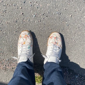 Maison Margiela sneakers med färgstänk - Unika vita sneakers från Maison Margiela med coola färgstänk i rött, orange, grönt och svart. Skorna har grå snörning, gummisula och är tillverkade i skinn. Perfekt för dig som vill sticka ut med en konstnärlig vibe.