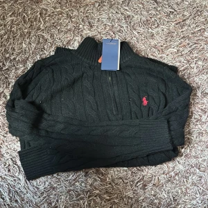 Svart stickad polotröja Polo Ralph Lauren - Svart polotröja från Polo Ralph Lauren i storlek M. Tröjan är kabelstickad med halv dragkedja och har den klassiska röda Polo-loggan broderad på bröstet. Hög krage och ribbade muddar ger en snygg och chill look. Perfekt för dig som gillar stilren design.