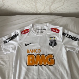 Vit Santos FC Nike fotbollströja - Santos FC matchtröja från Nike i vitt Dri-FIT-material. Klassisk design med klubbmärke, svarta detaljer och stora sponsorloggor i orange och svart. Nummer 11 på ryggen. Perfekt för dig som älskar brasiliansk fotboll och vill sticka ut på planen.