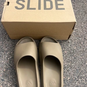 Adidas Yeezy Slide  - Säljer ett par Adidas Yeezy Slide i beige färg. Slidesen har en chunky, räfflad sula och är tillverkade i syntetmaterial för maximal komfort. Perfekta för chill dagar och har en stilren, minimalistisk look med öppen tå och bred rem. 44 1/2. Säljer billigt för vill bli av med dom, först till kvarn!