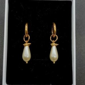 Guldiga örhängen med pärldroppar - Snygga örhängen med guldfärgade ringar och droppformade vita pärlor som hänger ner. Designen är elegant med en liten stjärndetalj ovanför pärlan. Perfekta för dig som vill ha en classy vibe och gillar att sticka ut med accessoarer.