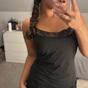 Svart linnetopp med spetsdetalj - Säljer en svart linnetopp med tunna axelband och snygg spets upptill vid urringningen. Toppen är figurnära och har en enkel, stilren look som passar perfekt till jeans eller kjol. Materialet känns mjukt och bekvämt mot huden. Även fast den sitter lite lösare på kroppen känns det fortfarande stylish. 