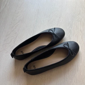 Svarta ballerinaskor med rosett - Stilrena svarta ballerinaskor från H&M med rund tå och dekorativ rosett framtill. Skorna har en platt sula och är tillverkade i ett syntetmaterial med mjuk innersula för extra komfort. Perfekta för en enkel och klassisk look.