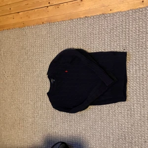Mörkblå stickad tröja Polo Ralph Lauren - Mörkblå stickad tröja från Polo Ralph Lauren med klassiskt kabelstickat mönster och röd broderad logga på bröstet. Tröjan har rund halsringning och ribbade muddar vid ärmslut och nederkant. Bara att höra av sig med frågor!