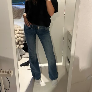 Blå bootcut jeans från Gina - Säljer ett par klassiska blå jeans från Gina med bootcut passform. Jeansen har låg midja, fem fickor och snygga detaljer med knappar på bakfickorna. Perfekt för dig som gillar en avslappnad och tidlös stil.