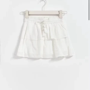 Vit volangkjol från Gina Tricot - Supersöt vit kortkjol från Gina Tricot med volanger och dekorativt snörning i midjan med pärlor och tofsar. Kjolen är gjord i mjukt bomullstyg och har en elastisk midja för skön passform. Storlek 146/152 men passar som xxs/xs💞 Aldrig använd