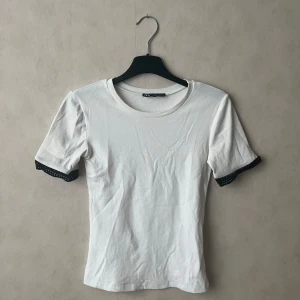 Vit t-shirt  - Vit t-shirt från Zara med rund halsringning och svarta spetsdetaljer längst ut på ärmarna. T-shirten har en tajt passform och perfekt för en clean och stilren look. Endast testad.
