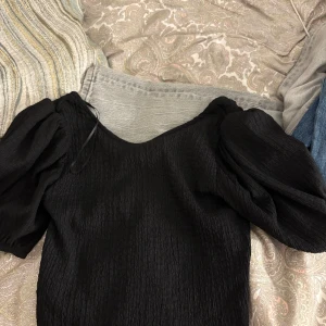 Svart blus med puffärmar - Supersnygg svart blus med puffiga korta ärmar och lätt skrynklig struktur. V-ringad halsringning och en trendig, elegant look som passar perfekt till jeans eller kjol. Toppens material känns lätt och luftig.