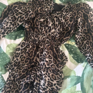 Leopardmönstrad kortklänning med volanger - Säljer en snygg kort klänning med leopardmönster i beige och svart. Klänningen har långa puffärmar, smock i midjan och volanger nedtill för extra attityd. Materialet är lätt och känns skönt mot huden, perfekt för dig som gillar att sticka ut.