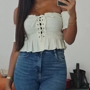 Offshoulder vit croppad topp med snörning - Supersöt offshoulder topp i vitt med smockad överdel, snörning framtill och volangkant nertill. Croppad modell som sitter tajt över bysten och har korta ärmar. Perfekt till höga jeans för en trendig look.