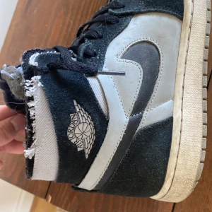 Nike Air Jordan 1 High svart/grå - Snygga Nike Air Jordan 1 High sneakers i svart och grått med klassisk vinge-logga på sidan. Skorna har högt skaft, snörning och en robust sula. Ovandelen är i mocka och skinn, med slitna detaljer för en cool, streetig vibe. Ny pris 1750