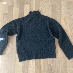 Mörkgrå stickad polotröja - Mysig mörkgrå stickad polotröja med hög krage och långa ärmar. Tröjan har breda ribbade muddar vid ärmslut och nederkant, perfekt för kalla dagar. Enkel och stilren design som passar till det mesta.