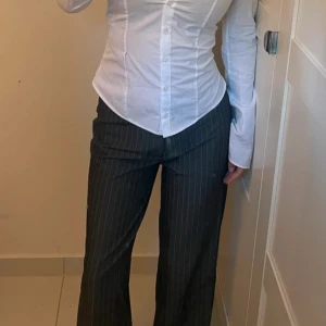 Randiga kostymbyxor från Madlady - Snygga grå kostymbyxor från Madlady med tunna vita ränder. Byxorna har mid/low midja, raka ben och bälteshällor. Perfekta för en clean och stilren look. Materialet känns mjukt och följsamt.  Jag har dock sytt om längden på byxorna så att de passar mig som är 158 cm