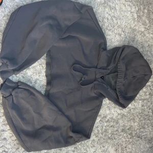 Mörkgrå croppad hoodie med snörning - Säljer en mörkgrå croppad hoodie med huva och snörning framtill. Tröjan har långa ärmar med ribbade muddar och är tillverkad i mjukt bomullsmaterial. Perfekt för en chill och avslappnad stil.