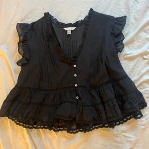 Svart volangblus från H&M - Supersöt svart blus från H&M med volanger på ärmar och nederkant, v-ringning med små knappar framtill och spetsdetaljer. Luftig modell. Aldrig använt 💕