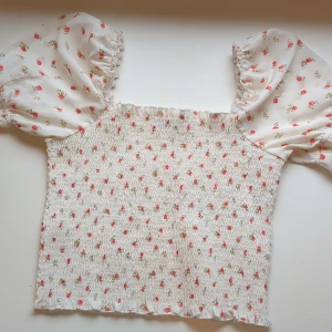 Blommig puffärm blus från NA-KD - Supersöt blommig blus från NA-KD med vita och rosa toner. Toppen har smockad kropp, volangkant nertill och puffiga korta ärmar i transparent tyg. Perfekta för sensommaren och skolstarten🫶