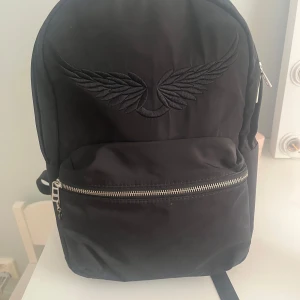 Svart ryggsäck Zadig & Voltaire - Cool svart ryggsäck från Zadig & voltarie med broderade vingar på framsidan och logga på axelremmarna. Rymligt huvudfack, framficka med drag och innerficka med etikett❤️ Ryggsäcken är rymlig så perfekt för skolan💖Ny pris 1700😇Pris går att diskutera💓