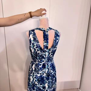 Blommig blåvit ärmlös klänning - Snygg ärmlös klänning med v-ringning och blommigt mönster i blått och vitt. Klänningen har en cool öppen rygg med två breda band och dragkedja bak. Passformen är normal och tyget känns som en lite tjockare bomull eller polyester. Perfekt för dig som gillar färg och mönster!