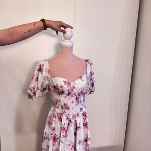 Vit blommig långklänning med puffärm - Romantisk långklänning i vit med rosa och lila blommor, puffärmar och markerad midja. Klänningen har ett sött sweetheart-utseende fram och en öppen rygg med dekorativ rem. Materialet är lätt och har en subtil struktur som ger extra känsla.