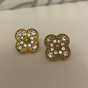 Guldiga örhängen med glittriga stenar - Supersnygga örhängen i guldton med fyrklöverform och glittrande vita stenar. De har små pärldetaljer runt kanterna och en cool, lyxig vibe som verkligen sticker ut. Perfekta för att ge din look lite extra bling och personlighet.