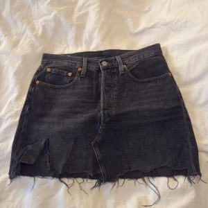 Svart jeanskjol från Levi's med fransig kant - Cool svart jeanskjol från Levi's med rå, fransig kant och slitna detaljer. Klassisk femficksmodell med knapp och dragkedja framtill samt Levi's-lapp bak i midjan. Perfekt för dig som gillar en edgy och avslappnad stil. (W:27)