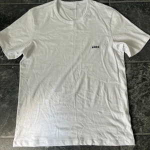 Vit BOSS t-shirt med broderad logga - Clean vit t-shirt från BOSS med svart broderad logga på bröstet. Klassisk rund hals och korta ärmar, tillverkad i mjuk bomull som känns skön mot huden. Perfekt basic med stilren vibe och enkel passform. Oanvänd, priset kan diskuteras vid snabb affär.