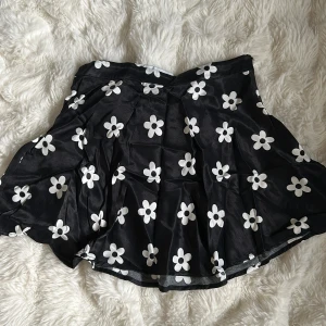 Svart blommig kortkjol från Bershka - Supersöt svart kortkjol från Bershka med vita blommor. Kjolen är lätt glansig. Stängs med dragkedja i sidan.