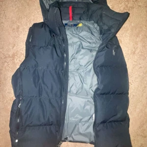 Svart dunväst från Polo Ralph Lauren - Snygg svart dunväst från Polo Ralph Lauren med huva och quiltad design. Västen har dragkedja framtill, praktiska fickor med dragkedja och en diskret logga på bröstet. Insidan är grå och materialet känns slitstarkt och varmt, perfekt för kyliga dagar.