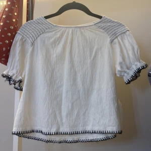 Vit blus med svarta detaljer från Zara - Superfin vit blus från Zara med svarta broderade detaljer längs ärmslut och nederkant. Blusen har korta puffärmar och rynkade partier vid axlarna. Tillverkad i ett lätt och luftigt bomullstyg, perfekt för en fräsch och stilren look.