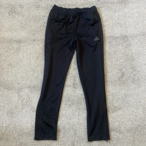 Svarta träningsbyxor från Adidas - Svarta träningsbyxor från Adidas med diskret logga på låret. Byxorna har elastisk midja och är tillverkade i ett lätt och stretchigt syntetmaterial, perfekt för träning eller chill. Stl 152/11-12 år 